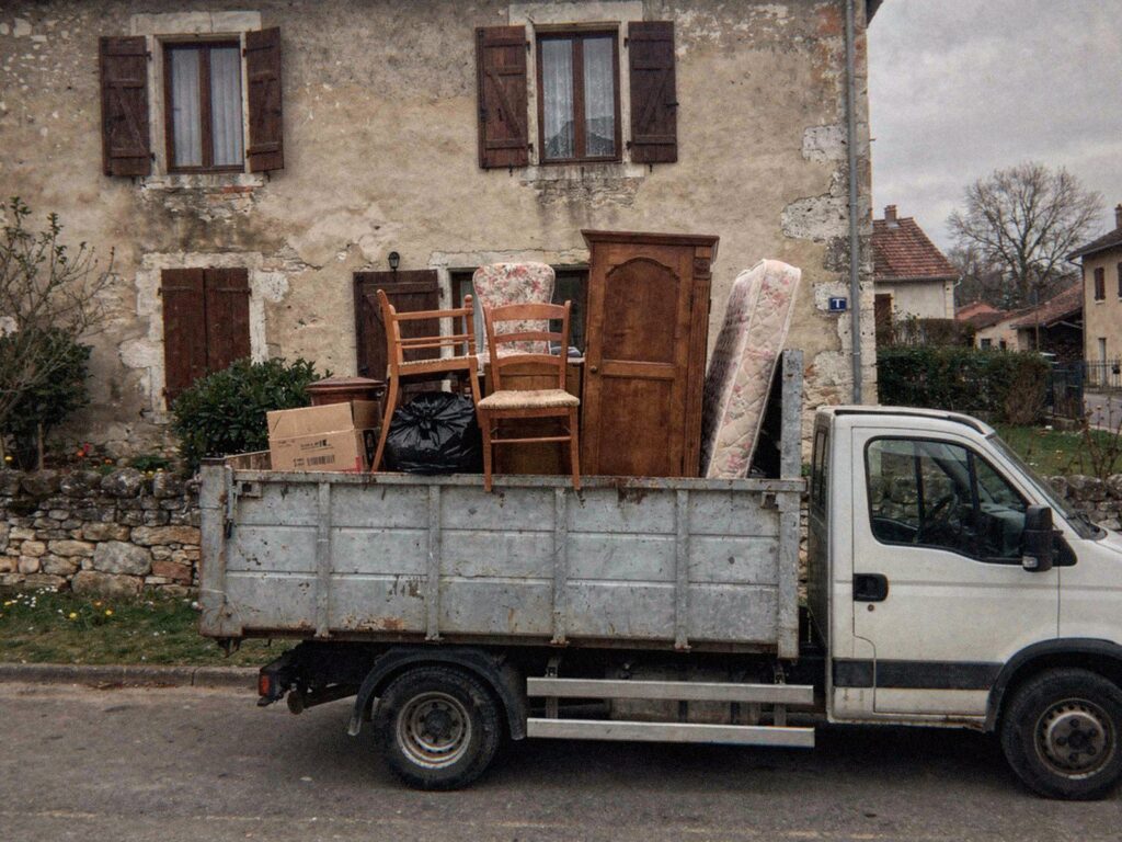 Camion de MJ Débarras chargé de chaises, d'une armoire et d'un matelas devant une maison de l'Ain